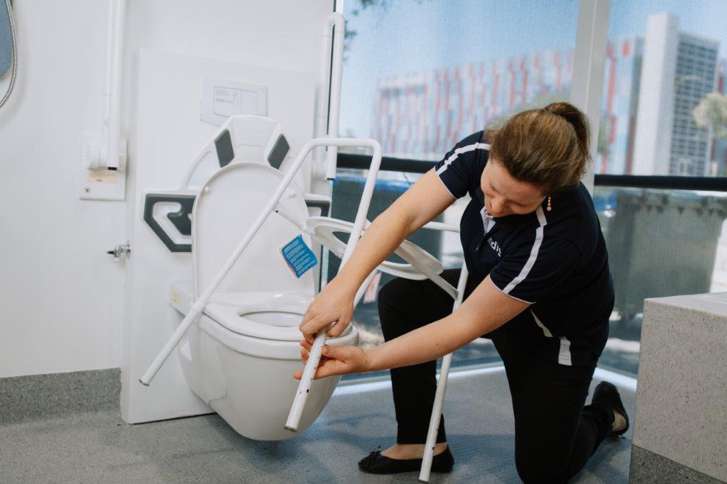 Female Indigo OT kneeling to adjust toilet frame in bathroom display